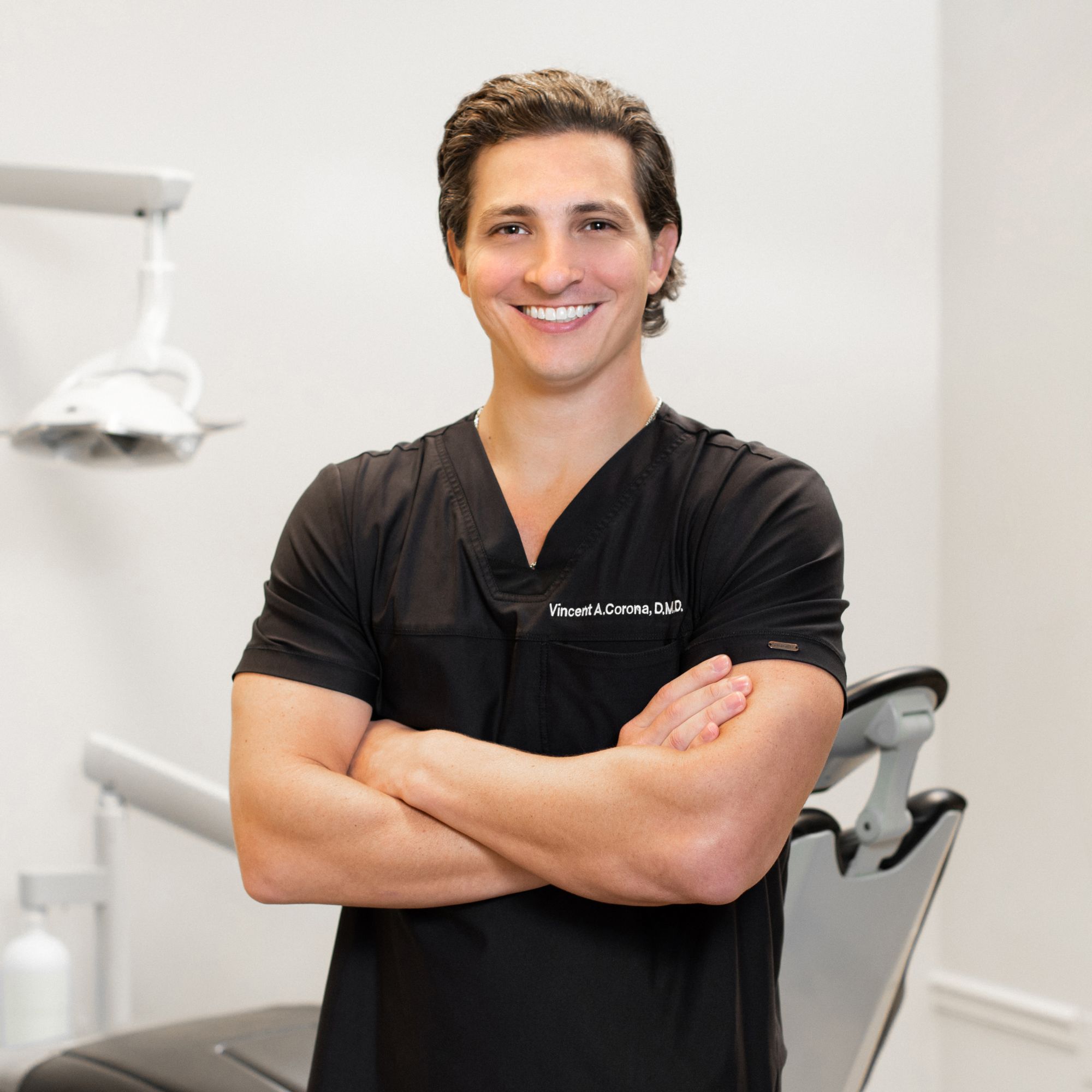 A male dental professional standing confidently with arms crossed in front of a dental office setting.
