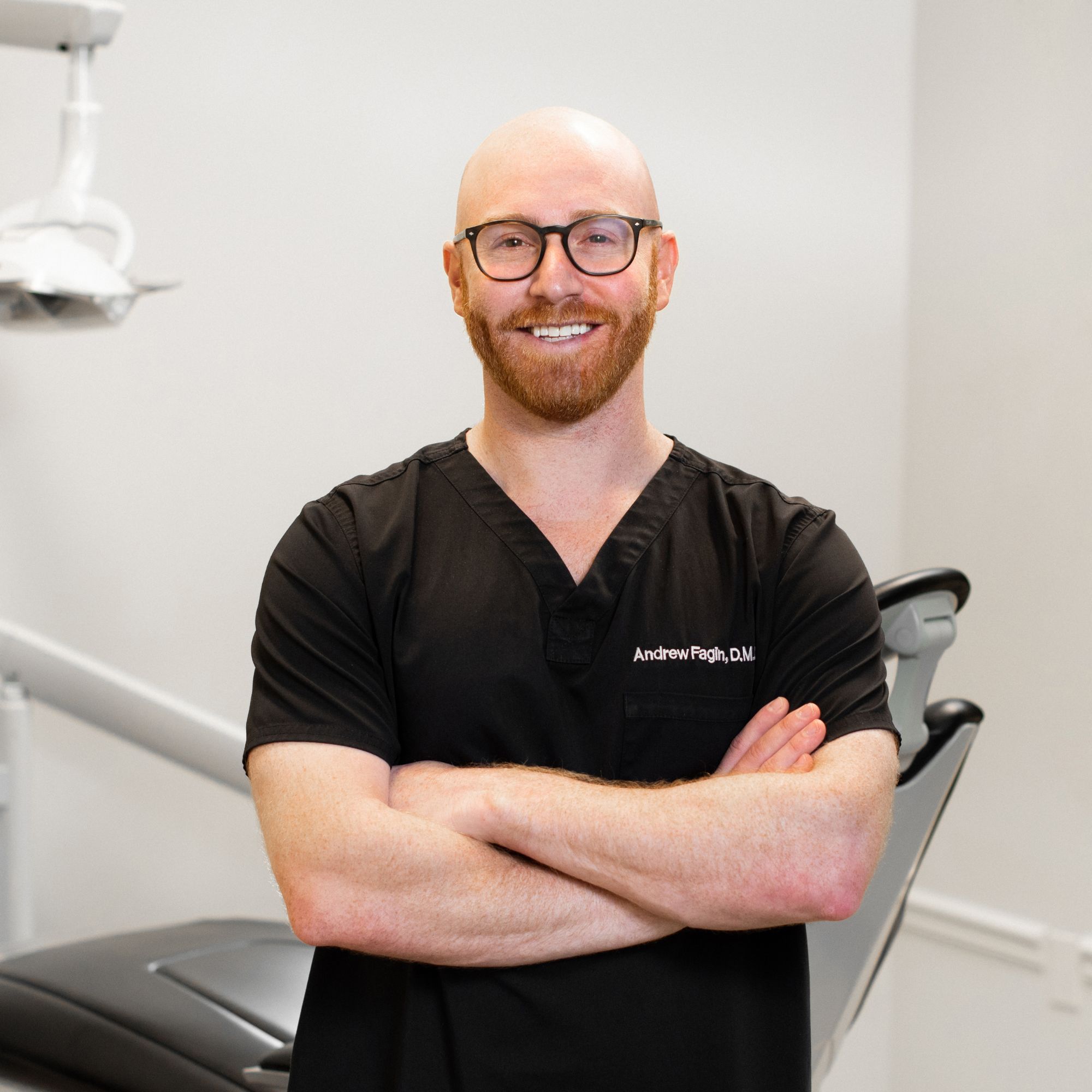 The image shows a man standing confidently with his arms crossed in front of him, smiling at the camera. He is wearing glasses, a dark shirt, and has short hair. Behind him is dental equipment, indicating he may be a dentist or in a dental setting.