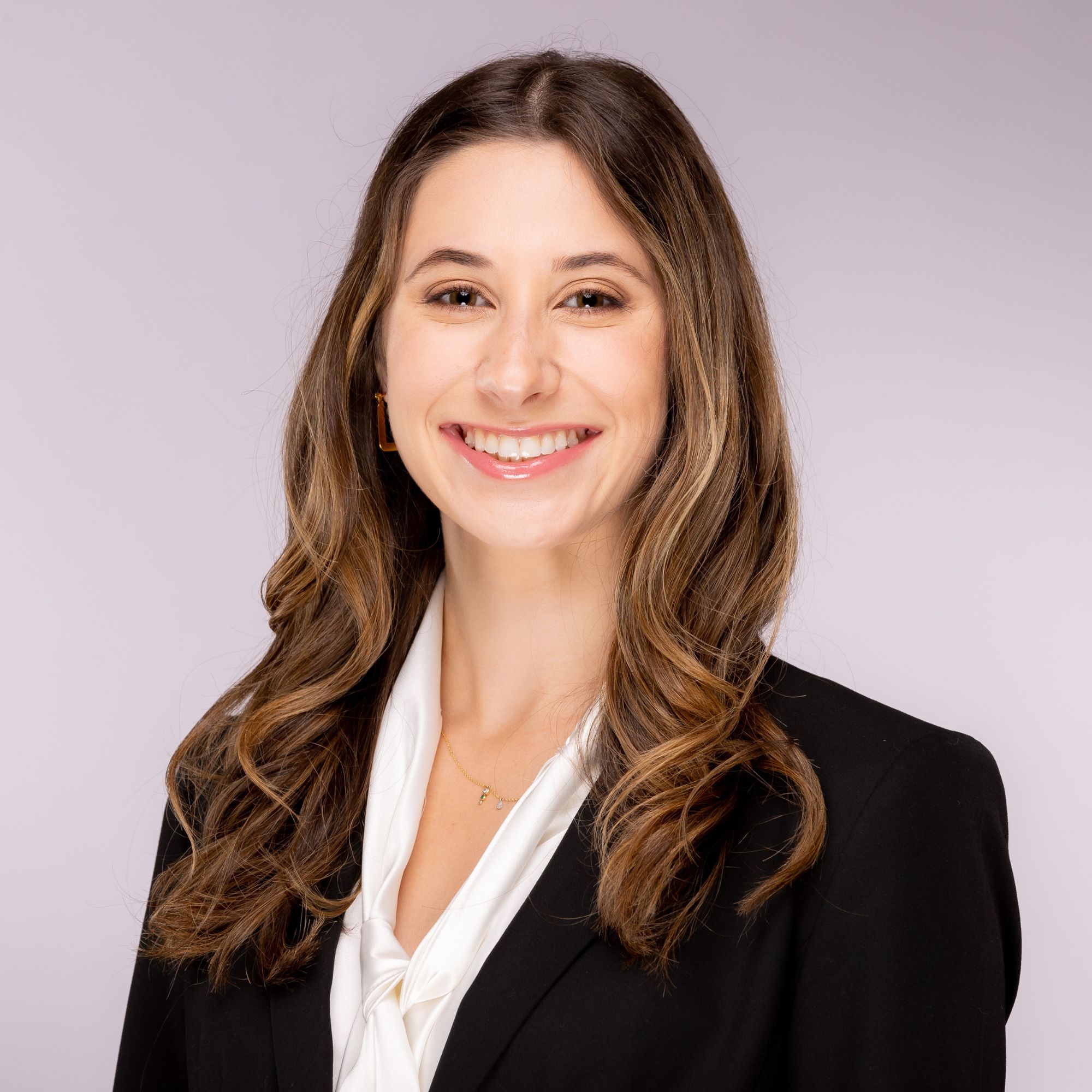 The image shows a woman smiling at the camera, wearing a dark blazer over a lighter top with long hair, standing against a neutral background.