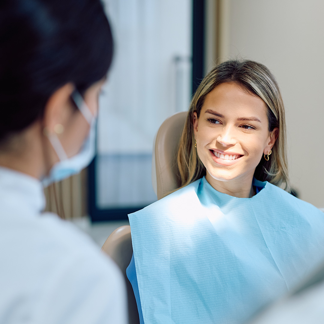 The image shows a woman in a dental office setting, sitting in a dentist s chair with a smile on her face, receiving dental care from a professional wearing a surgical mask and stethoscope.