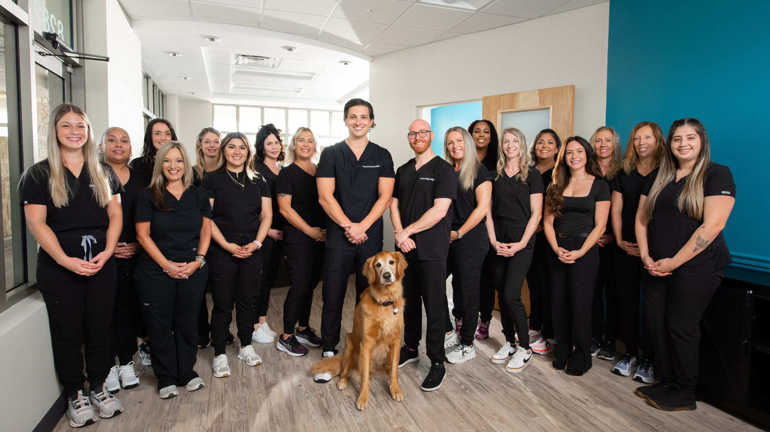 The image shows a group of people posing for a photo, with a man standing at the center and a dog on the floor behind them. They are indoors, likely in an office setting, with some individuals wearing uniforms that suggest they may be dental professionals.
