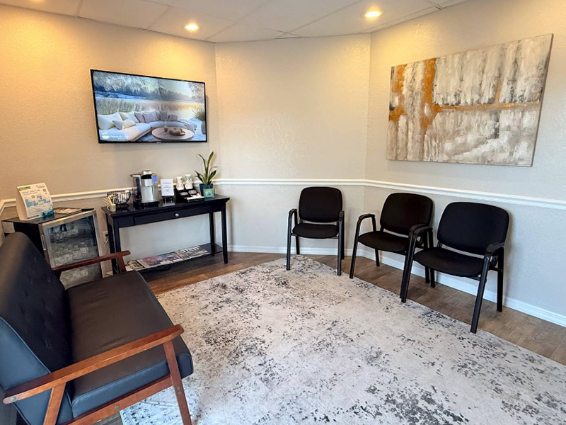 The image depicts an interior view of a waiting room with a modern design featuring a large window, white walls, a black chair, a reception desk, and a painting on the wall.