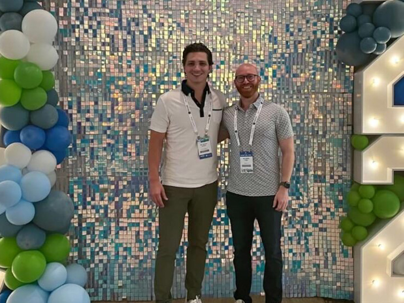 Two men standing side by side at an event with balloon backdrop featuring numbers and a colorful mosaic wall, posing for a photo.