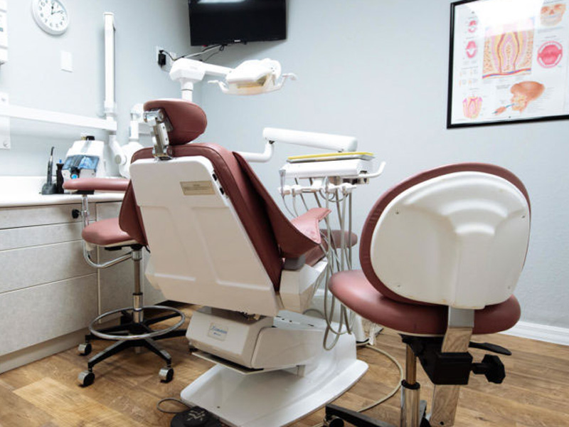 The image shows a dental office interior with a dental chair, a dental table, and dental equipment, including a dental mirror, suction unit, and other tools.