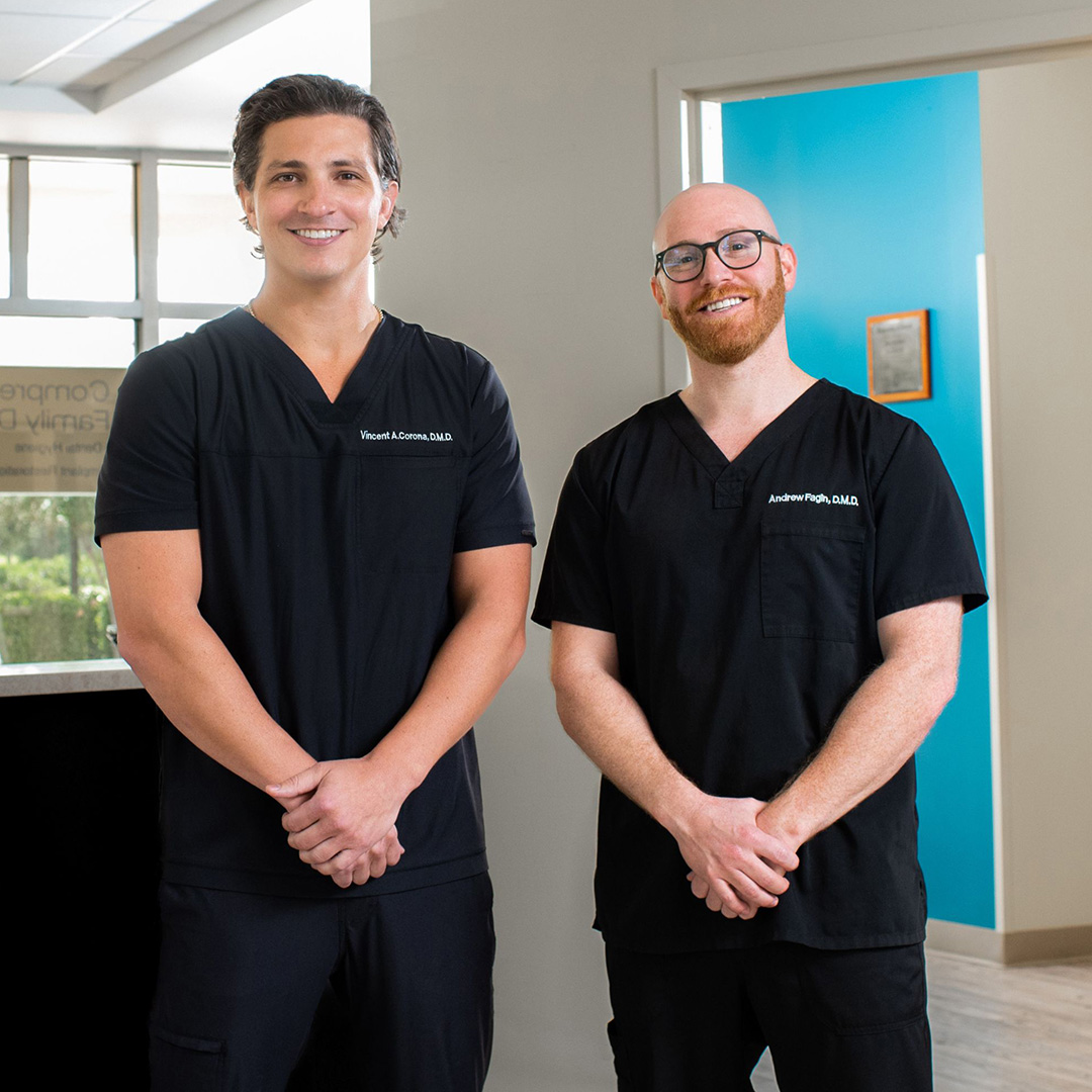 Two individuals standing side by side in an indoor setting, both wearing scrubs with name tags, posing for a photo in front of a wall-mounted sign with text.