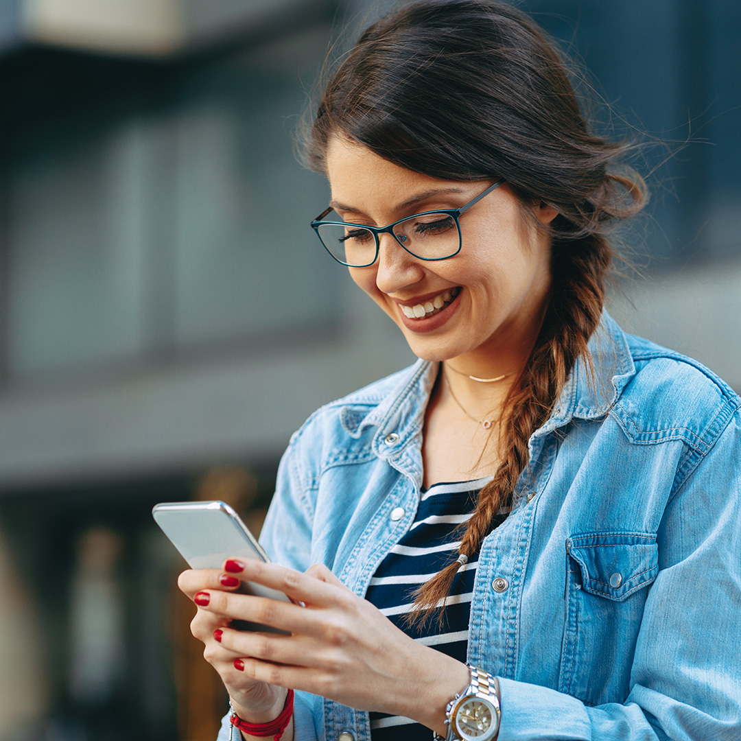 A woman with glasses and braided hair is using her phone while standing on a street.