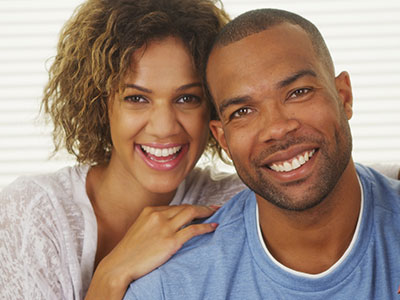A man and woman pose happily together, smiling at the camera with the man wearing a blue shirt and the woman in a white top.