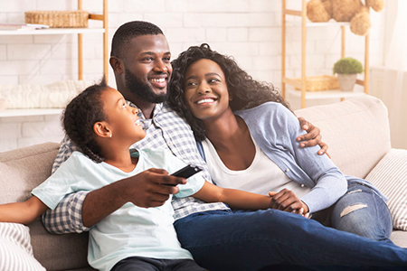 A family of four enjoying time together on a couch, with an adult male, female, and two children smiling at the camera.