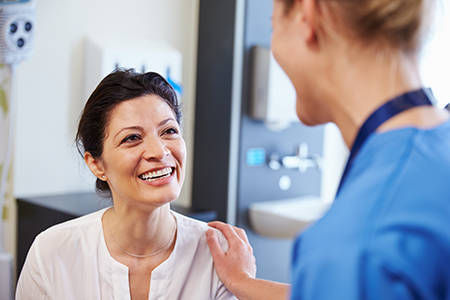The image shows a woman with short hair smiling at another woman who appears to be a medical professional, possibly a nurse or doctor, in an indoor setting that looks like a hospital or medical office.