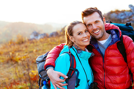Two people, a man and woman, are smiling and embracing outdoors during daylight. They appear to be hikers, with the man wearing a backpack and both dressed for cold weather.