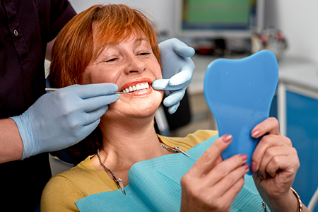 A woman in a dental office holding up a blue toothbrush with a smile on her face while wearing a surgical mask, surrounded by dental equipment and a professional in a lab coat.