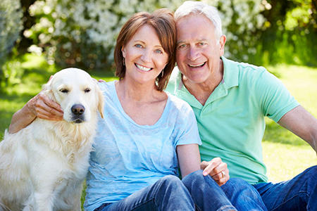 In the image, there are two people and a dog sitting on grass. The man is wearing a green shirt and the woman is wearing a blue top. They are both smiling and looking at the camera.
