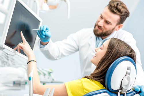 A man and woman are seated at a computer screen with medical equipment behind them  the man appears to be demonstrating something to the woman, who is wearing headphones and looking intently at the screen.