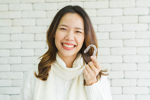 A woman with a smile, holding a white object, against a brick wall background.