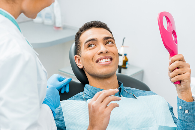 The image shows a person sitting in a dental chair with a smiling expression, holding a pink object, possibly an oral care device, while being attended by a dental professional who appears to be preparing them for a dental procedure.
