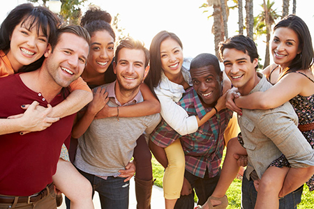 The image depicts a group of people posing together outdoors during daylight with bright lighting and clear skies, smiling and embracing each other.