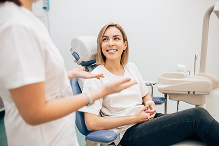 The image depicts a woman sitting in a dental chair with a smile on her face, surrounded by dental professionals who appear to be engaged in conversation or examination.