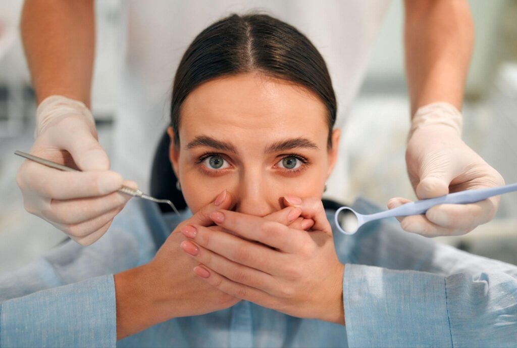 A nervous woman covers her mouth while a dentist in gloves holds dental tools, representing dental anxiety and fear of routine oral health checkups