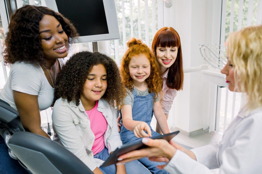 Two mothers and their daughters smiling and talking to a dentist at a family dental clinic, showing a relaxed and friendly environment for pediatric and family dentistry in Sarasota, FL.