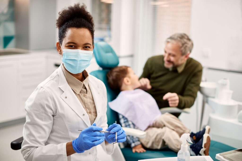 Female dentist in Sarasota, FL smiling at camera while a young child sits in the dental chair with a parent, showing friendly family dental care.